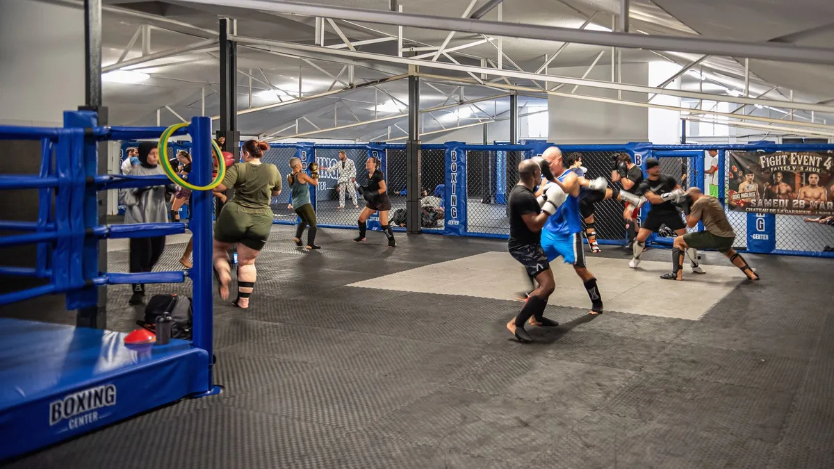 La grande salle du Boxing Center Toulouse États-Unis : le ring au premier plan, la cage de MMA au fond, un cours en séance.
